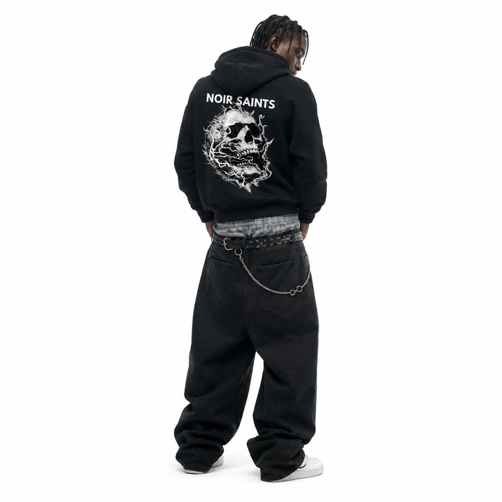 A model viewed from behind wearing a black hoodie with a large white skull graphic and the text 'NOIR SAINTS' on the back, styled with baggy black jeans, a chain belt, and white sneakers against a plain white background.
