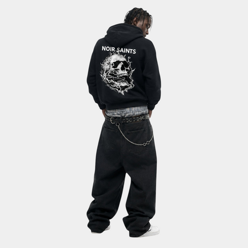 A model viewed from behind wearing a black hoodie with a large white skull graphic and the text 'NOIR SAINTS' on the back, styled with baggy black jeans, a chain belt, and white sneakers against a plain white background.